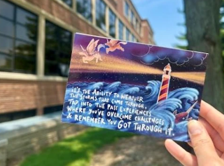A person holding up a colorful postcard outside. The postcard shows a scene of a lighthouse amidst waves. The text reads 